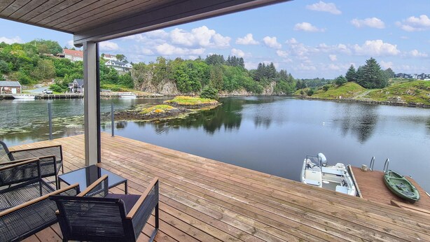 A wooden deck with outdoor seating, overlooking a calm river with boats and greenery. - 5 bedroom gorgeous home in Bremnes (Bremnes)