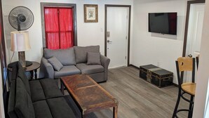 Ein Wohnzimmer mit einer Couch, einem Holztisch, einer Stehlampe, einem Ventilator, einem Fernseher und einem Koffer.