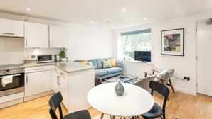 A modern kitchen with white cabinets, a microwave, and a stove. There is a dining area with a round table and chairs, and a blue sofa with cushions. A TV is mounted on the wall, and a window provides natural light. - Shoreditch | 3 King Beds | Fast Wi-Fi | Balcony (London)