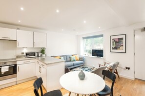 A modern kitchen with white cabinets, a microwave, and a stove. There is a dining area with a round table and chairs, and a blue sofa with cushions. A TV is mounted on the wall, and a window provides natural light. - Shoreditch | 3 King Beds | Fast Wi-Fi | Balcony (London)