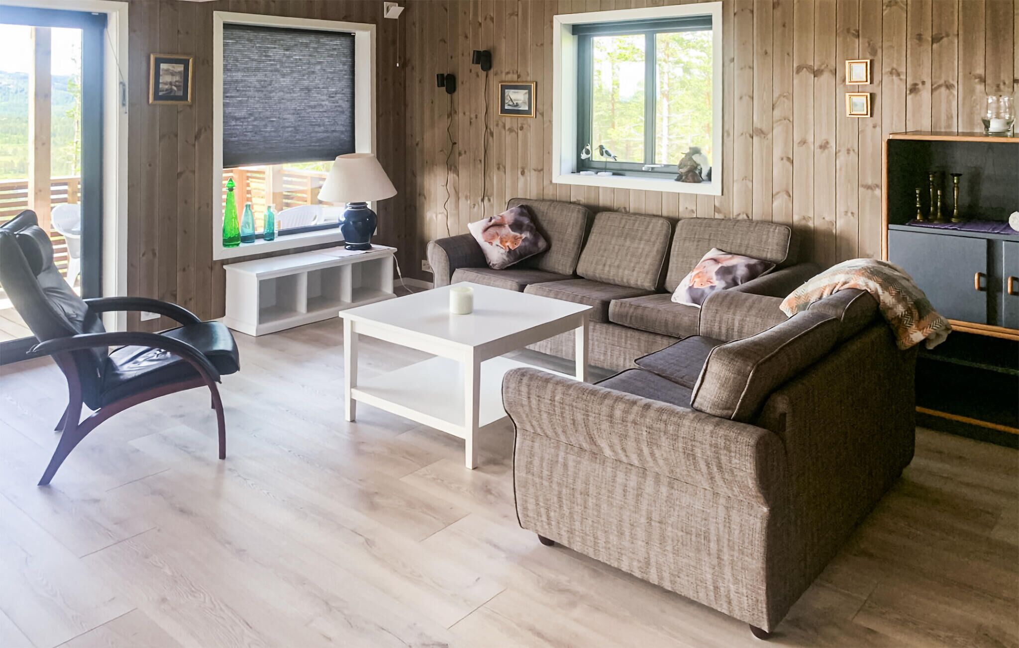 Un salon confortable avec un canapé, une table basse et un fauteuil. Il y a deux fenêtres avec des stores, une lampe sur une table d’appoint et une étagère avec des bouteilles.