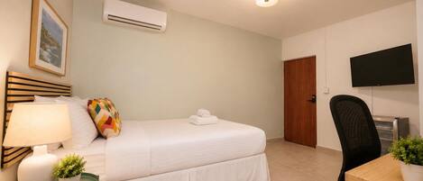 A hotel room with a bed, a desk, a chair, a TV, an air conditioner, and a framed picture on the wall.