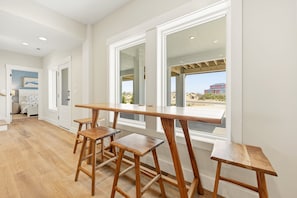 A spacious room with a large wooden table and three stools, large windows with a view, and a door leading to another room.