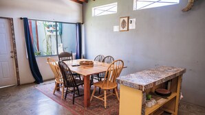 Een eetgedeelte met een houten tafel en stoelen, een klein plankje met servies en uitzicht buiten door een raam.