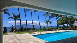 A pool area with a view of the ocean and mountains, surrounded by palm trees and a covered patio. - Lahaina Shores 527 – Relaxing Oceanfront Escape (Lahaina)
