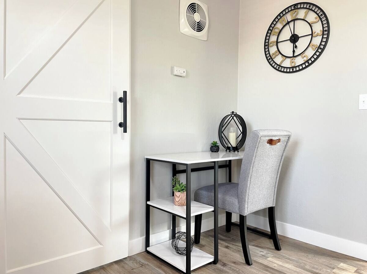 A room with a white door, a small table with a chair, a wall clock, and a fan.