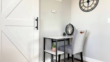 A room with a white door, a small table with a chair, a wall clock, and a fan.
