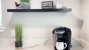 A coffee maker with a mug on a kitchen counter, a potted plant, and a shelf with a coffee-themed sign.