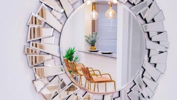 Espejo redondo con marco geométrico que refleja una habitación con una ventana, una planta y dos sillas.