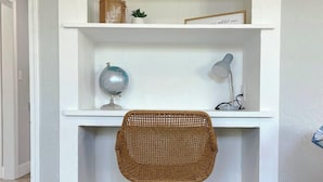 A corner workspace with a white desk, a wicker chair, a globe, a lamp, and decorative items on shelves.
