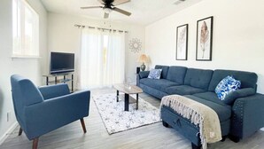 Una sala de estar con un sofá azul, un sillón azul, una mesa de centro, un ventilador de techo y un televisor.