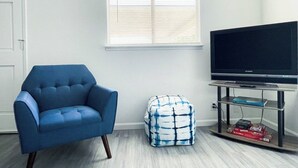 Una habitación con un sillón azul, un reposapiés blanco y azul, un televisor sobre un mueble bajo y una ventana con persianas.