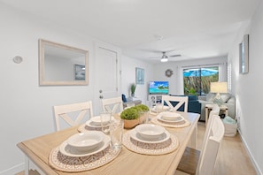 A dining area with a wooden table set for four, white chairs, a ceiling fan, and a television in the background.