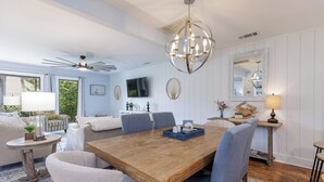 Un coin repas avec une table en bois, des chaises bleues et un lustre. Un téléviseur est fixé au mur et un ventilateur de plafond est installé.