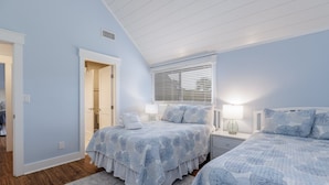 Une chambre avec deux lits, une fenêtre avec des stores, une table de chevet avec une lampe et une porte donnant sur une autre pièce.