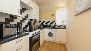 Une cuisine compacte avec des meubles blancs, des carreaux noirs et blancs, un four encastré, un four à micro-ondes, un lave-linge et un évier.
