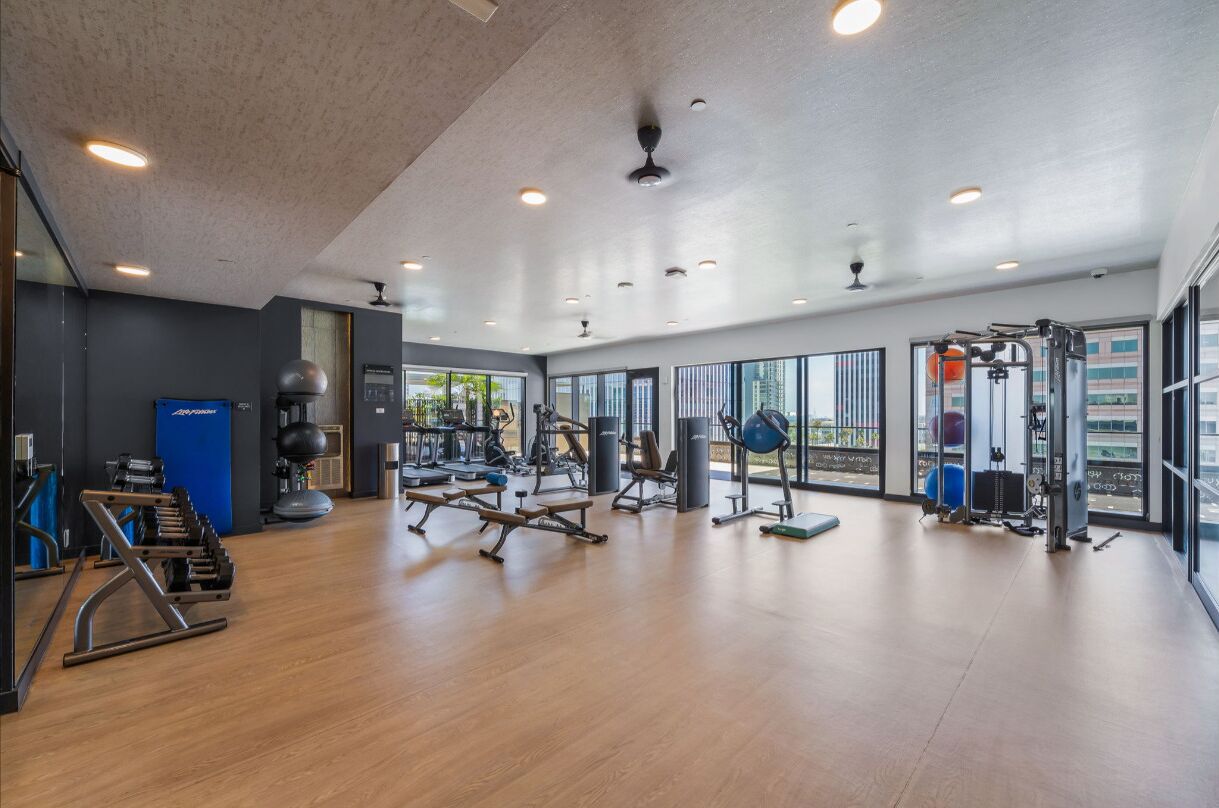 Un gimnasio espacioso con diverso equipo de ejercicio, ventanas amplias y un techo alto.