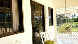 A room with two white plastic chairs with green cushions, a small wooden table, and a large window with a view of greenery and a hammock. - The Ranch Jamaica Eco Resort and Ranch (St. Catherine Parish)