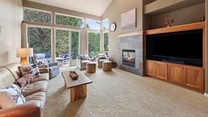 Un salon spacieux avec un canapé en cuir, des poufs, une table basse et une cheminée. Une grande fenêtre offre une vue sur les arbres, et une horloge est accrochée au mur.