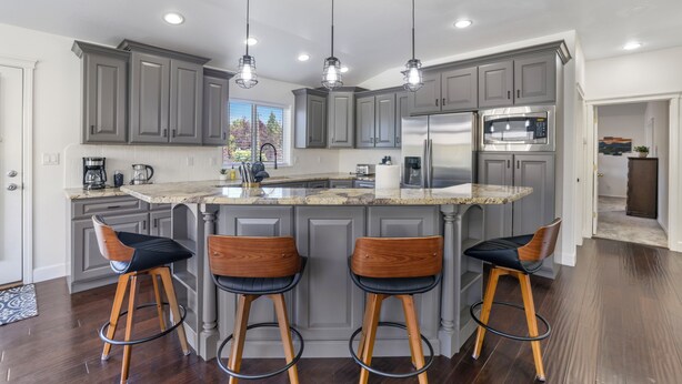 A modern kitchen with grey cabinets, a stainless steel refrigerator, a built-in microwave, and a granite countertop. There are three wooden bar stools with black cushions.