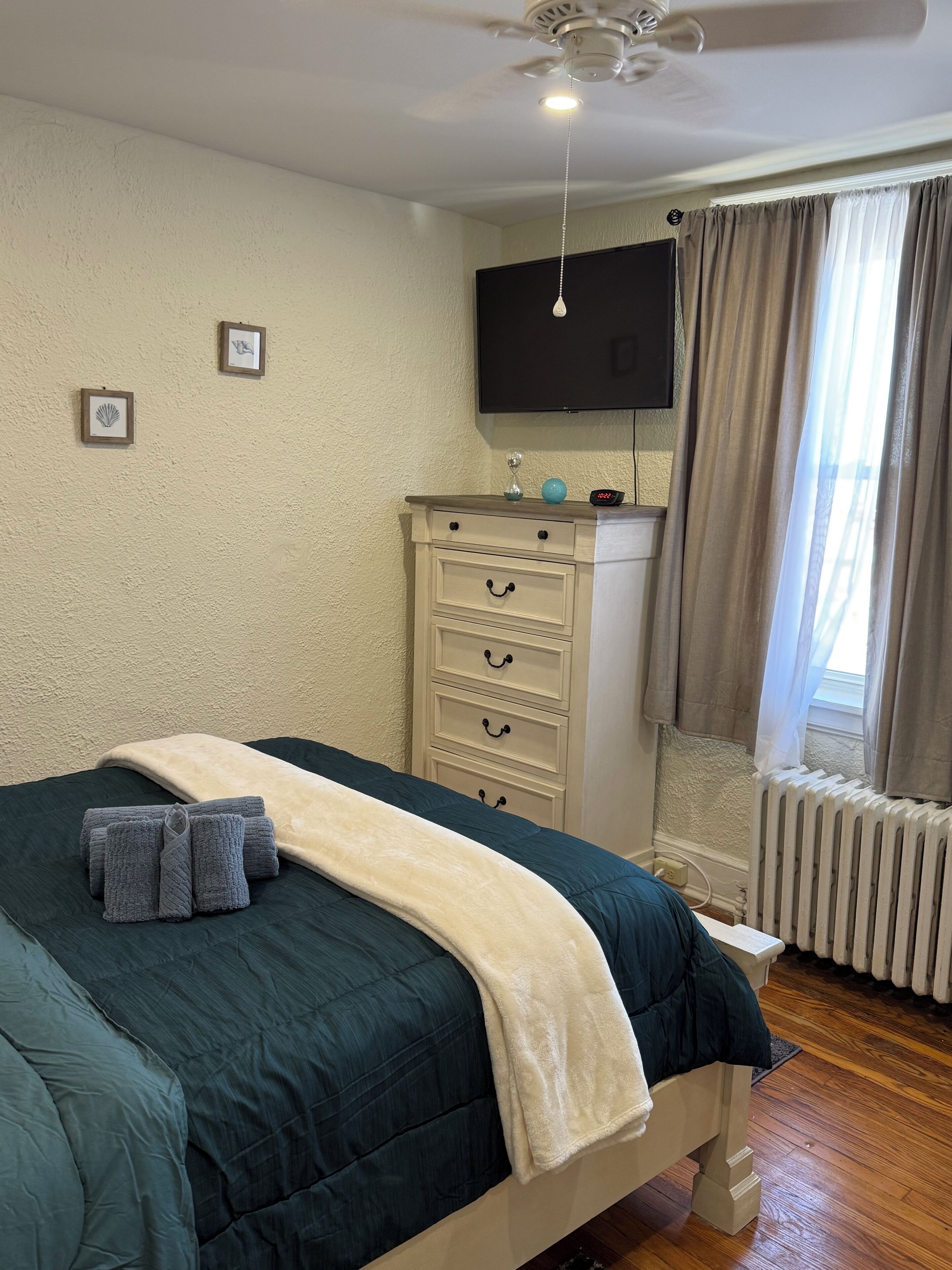 Ein Schlafzimmer mit Bett, Kommode, Fernseher und einem Fenster mit Vorhängen.