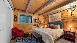A cozy log cabin bedroom with a large bed, a red chair, a wooden bench, and a window with a view of trees.