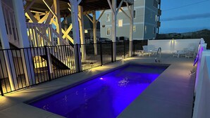 A rectangular swimming pool with blue lighting, surrounded by a white fence, and adjacent to a building with a covered deck.