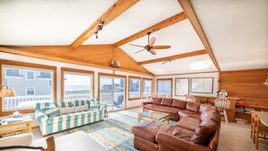 A spacious living room with a high ceiling, wooden beams, large windows, and a view of the sea. There are two sofas, a coffee table, and a TV.