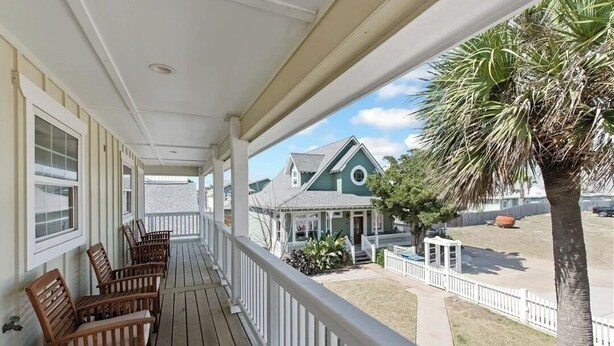 A wooden porch with white railings, wooden chairs, and a view of a green house with a white fence and a palm tree. - Pet Friendly, Close to the Beach, Golf Cart Access (Port Aransas)