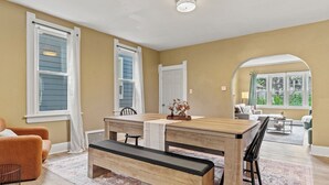 A dining area with a wooden table and bench, beige walls, and large windows. - Brady St Retreat Front House - Sleeps 8 & Pets (Milwaukee)