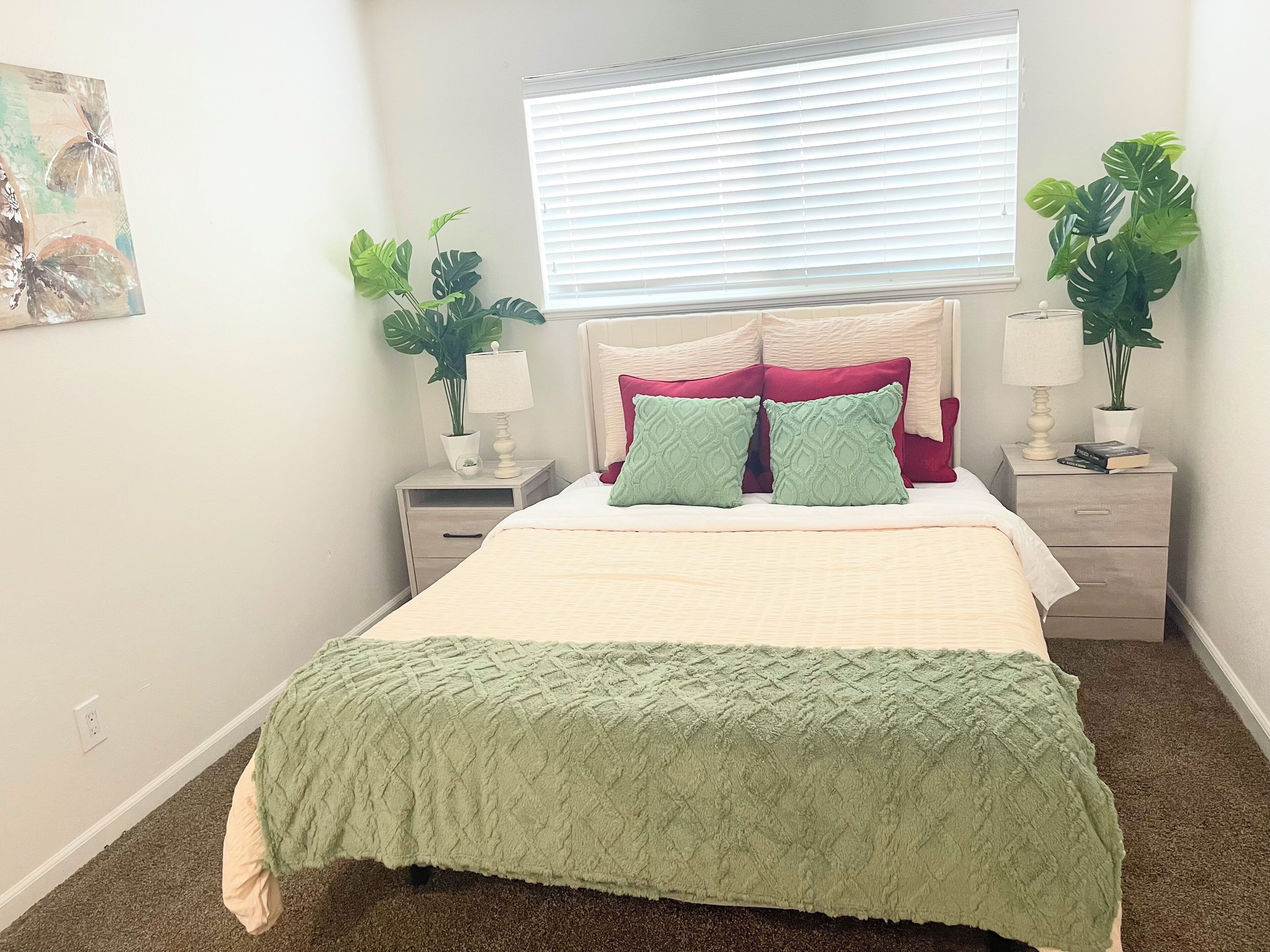A neatly arranged bedroom with a bed, two bedside tables, a window with blinds, and two potted plants.