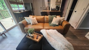 A modern living room with a leather sofa, black ottoman, and a fluffy white throw. A wooden tray with a plant and remote control is on the ottoman. - Charming 2-bedroom cabin in delightful New Market with AC (New Market)
