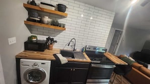 A compact kitchen with a washing machine, a microwave, and a stove. There are wooden shelves above the sink and a small plant on the counter. - Charming 2-bedroom cabin in delightful New Market with AC (New Market)