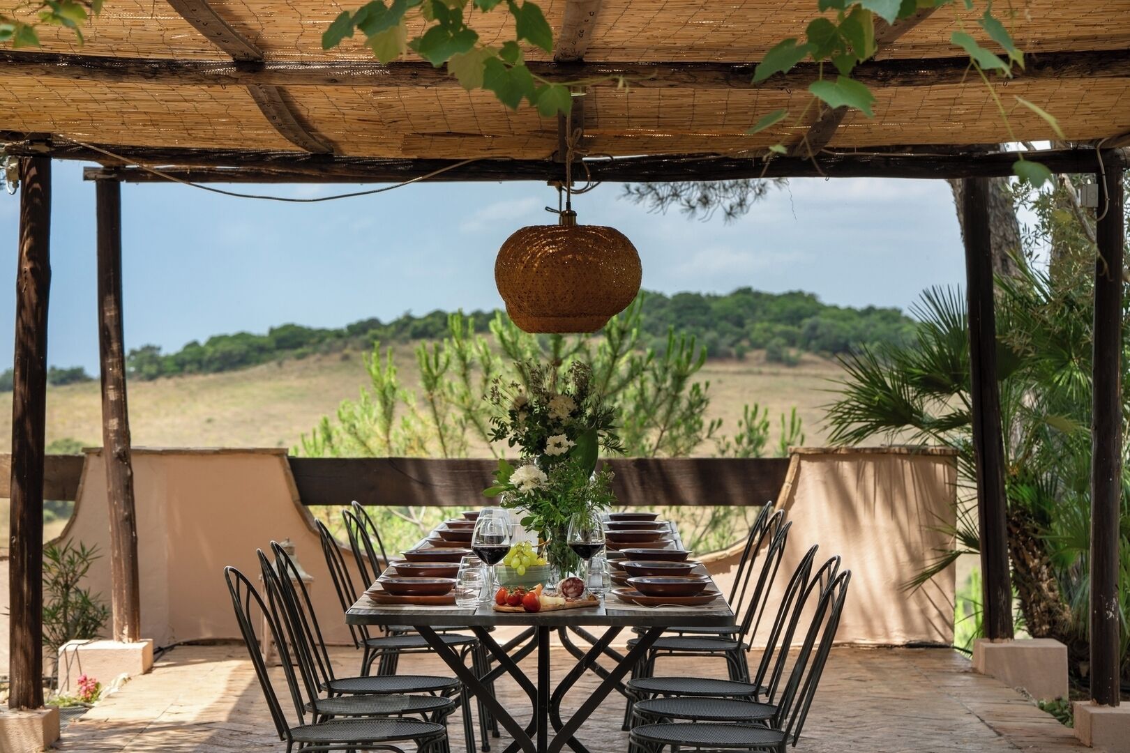An outdoor dining area with a table set for six, surrounded by chairs, under a wooden pergola with a hanging light fixture.