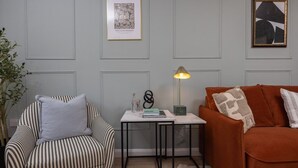 Un salón con un sillón a rayas, un sofá marrón, una mesita con una lámpara y cuadros en la pared.
