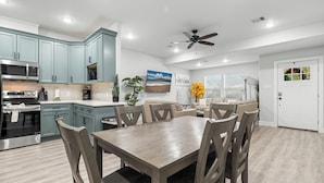 Una cocina moderna con armarios verde azulado, una zona de comedor con una mesa y sillas de madera, y un ventilador de techo.
