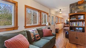 Una sala de estar acogedora con un sofá verde, estanterías de madera y grandes ventanales.