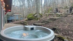 Un jacuzzi en un patio trasero con un muro de piedra, escalones de madera y un área boscosa al fondo.