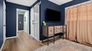 Habitació amb una paret blava fosca, un paviment de fusta, un televisor de pantalla plana muntat sobre una commode i una cortina beig.