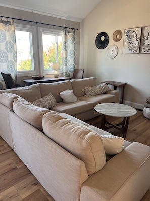 A living room with a beige sectional sofa, a round wooden coffee table, and a window with patterned curtains.