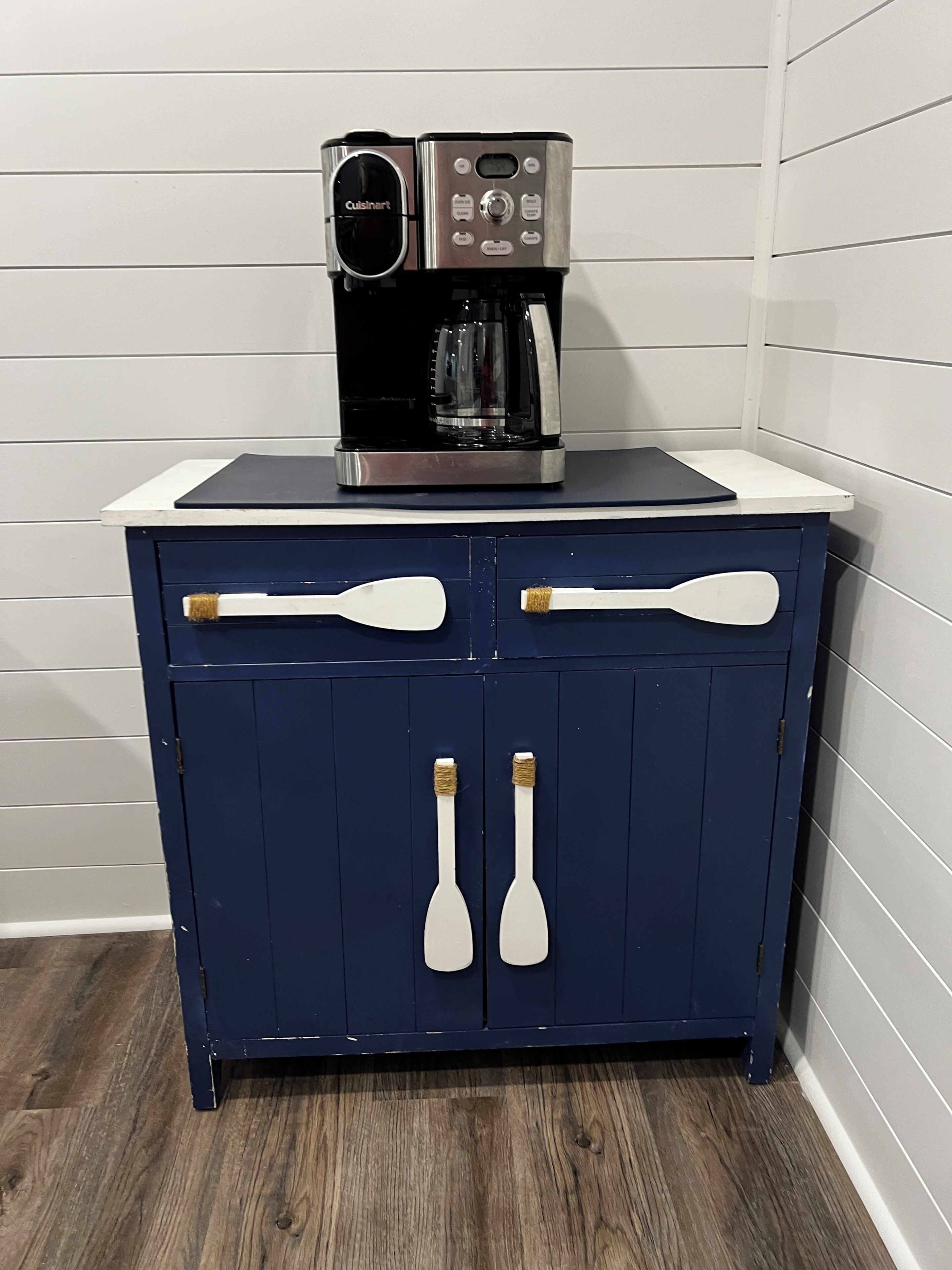 Eine Küche mit einer Kaffeemaschine auf einer Arbeitsfläche, einem blauen Schrank mit zwei Griffen und einer weißen Arbeitsplatte.