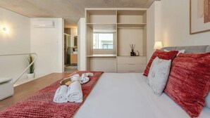 Una habitación de hotel con una cama, una colcha roja, toallas, una mesita de noche, una lámpara y un armario.