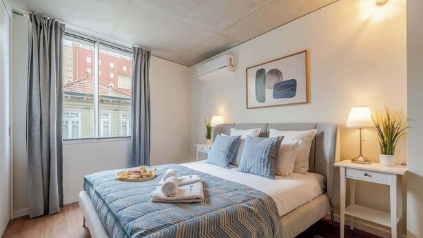 Een hotelkamer met een groot bed, een nachtkastje met een lamp, een raam met gordijnen en een lijst met een afbeelding aan de muur.