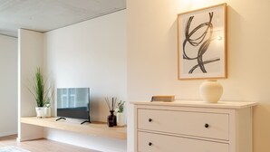 Een moderne woonkamer met een flatscreen televisie op een houten plank, een witte kast met lades, een lijst met abstracte kunst en een potplant.