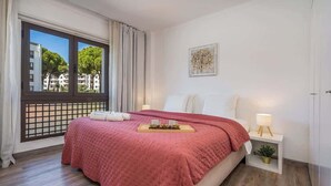Quarto com cama, janela com vista para edifícios e árvores, mesa de cabeceira com abajur e uma pequena planta.