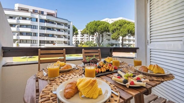 Varanda com mesa de café da manhã, incluindo croissants, frutas e suco, com vista para uma área residencial com árvores e edifícios.