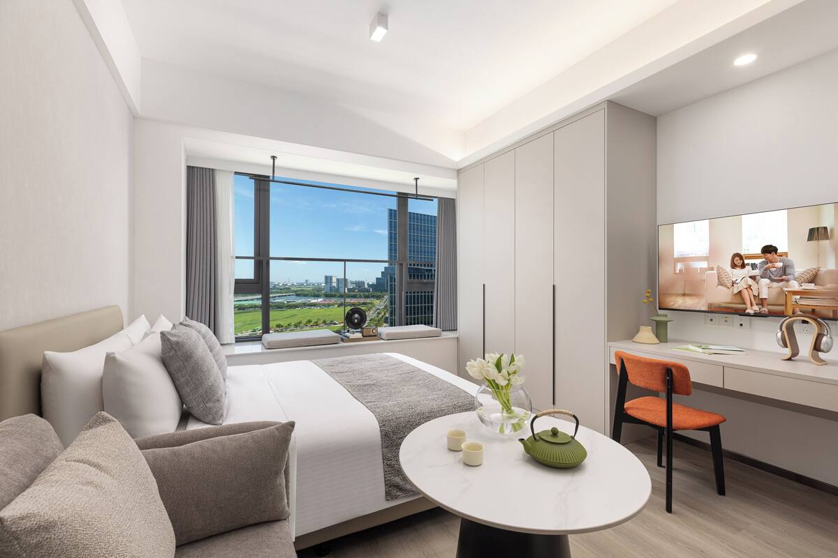 A modern hotel room with a large bed, a desk with a chair, a round table, and a large window offering a city view.