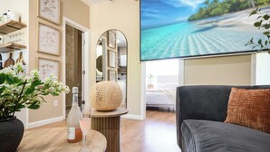 Un salon moderne avec un canapé gris, une table ronde en bois sur laquelle se trouvent une bouteille et un verre, et un grand écran diffusant une scène de plage.
