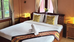 A neatly made bed with white linens, a decorative throw, and two pillows with text. There is a bedside table with a lamp, a wall clock, and a window with curtains. The room has wooden flooring and a wooden ceiling with exposed beams. - The Nest Pai Love Resort (Pai)
