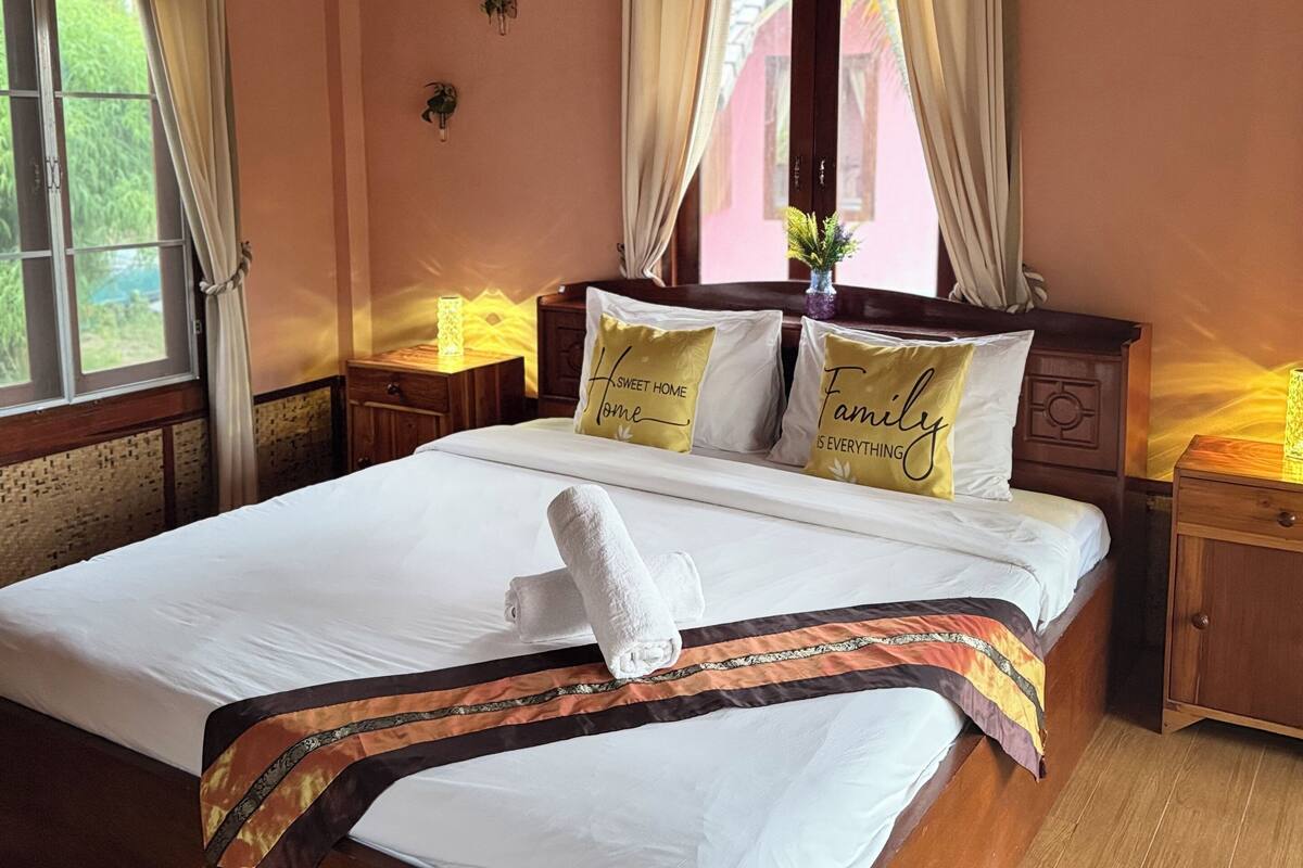 A neatly made bed with white linens, a decorative throw, and two pillows with text. There is a bedside table with a lamp, a wall clock, and a window with curtains. The room has wooden flooring and a wooden ceiling with exposed beams.